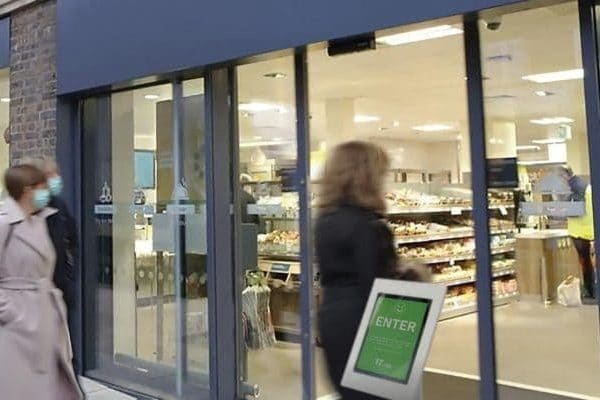 Bezoekers lopen een winkel binnen terwijl een digitale bezoekersteller met stoplichtfunctie via het scherm bij de ingang laat zien dat het maximale bezoekersaantal (in het kader van corona) nog niet is bereikt