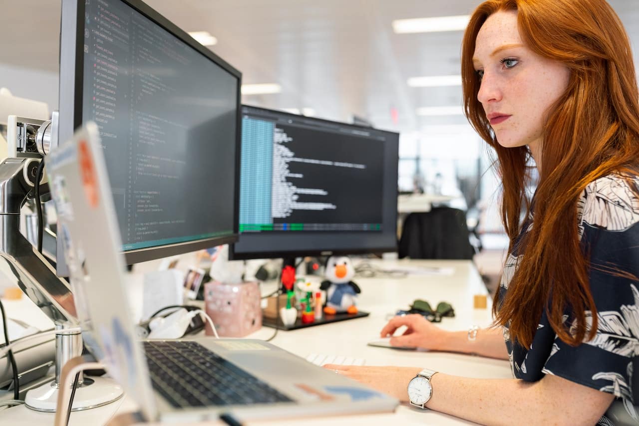 Een jonge vrouw is aan het programmeren achter meerdere beeldschermen